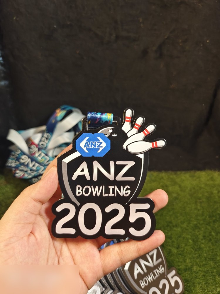 Medal ANZ Bowling 2025 diambil dekat dlm genggaman — jelas tunjuk bentuk perisai hitam, logo ANZ biru, pin bowling putih dan teks '2025' tebal. Bagus untuk tunjuk saiz sebenar berbanding tapak tangan.
