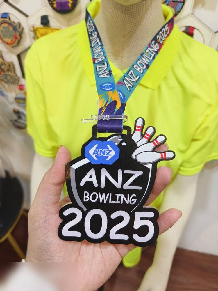 Medal ANZ Bowling 2025 — bowling korporat. Bentuk custom perisai dengan pin bowling dan bola timbul di sudut kanan atas. Warna hitam dengan logo ANZ biru. Teks besar 'ANZ BOWLING 2025'. Lanyard bercet