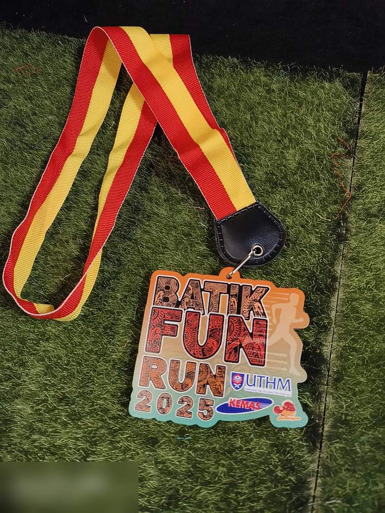 Medal Batik Fun Run 2025, anjuran UTHM dan KEMAS. Bentuk custom dgn teks besar bercorak BATIK sebenar di dalam huruf, warna oren dan coklat dgn silhouette pelari. Reben merah-kuning dgn tab kulit hita