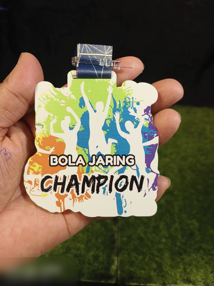 Medal 'BOLA JARING CHAMPION'. Bentuk custom dengan latar putih dan percikan cat warna-warni (hijau, biru, oren, ungu) serta silhouette pemain meraikan kemenangan. Reben biru gelap bercorak. Sesuai unt