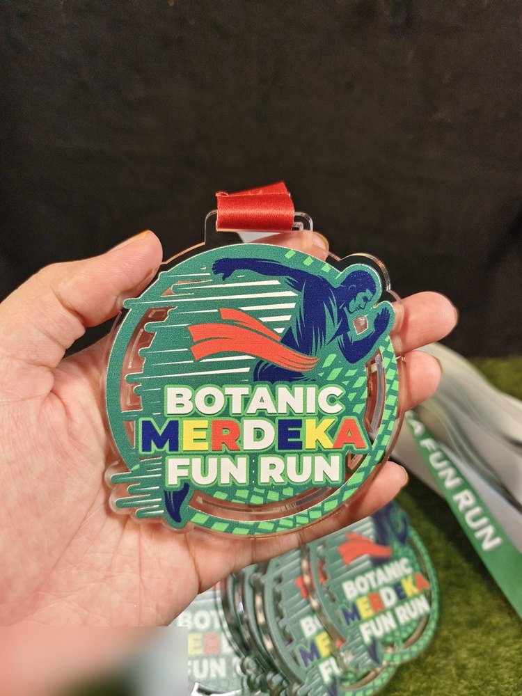 Medal Botanic Merdeka Fun Run. Bentuk bulat acrylic lut sinar dengan design hijau dan biru — pelari memakai jubah merah, garisan kelajuan dan trek berpetak. Teks 'BOTANIC MERDEKA FUN RUN'. Reben merah