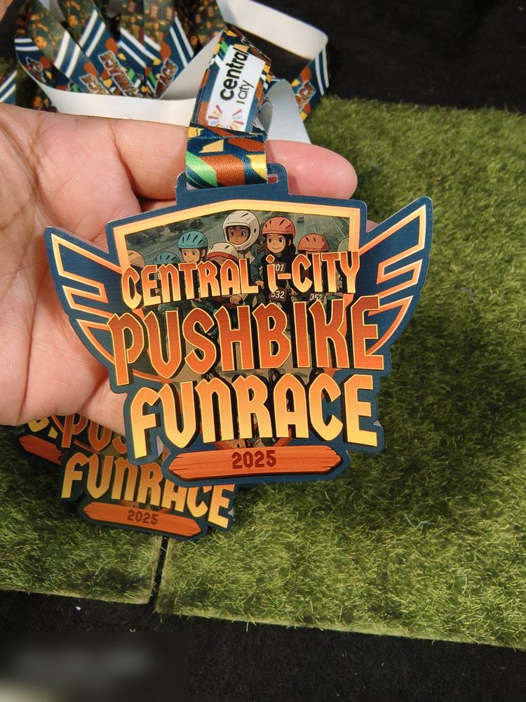 Medal Central i-City Pushbike Funrace 2025. Bentuk custom BERSAYAP, latar biru navy dgn teks oren-kuning tebal dan GAMBAR FOTO sebenar kanak-kanak berhelmet dimasukkan ke dalam design. Contoh cetakan 