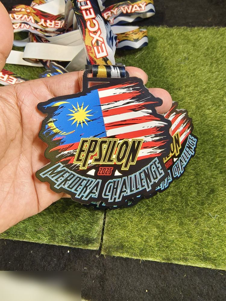 Medal Epsilon Merdeka Challenge 2025. Bentuk custom mengikut Jalur Gemilang berkoyak (torn flag effect) — biru, merah, putih dan kuning. Teks 'EPSILON MERDEKA CHALLENGE 2025'. Lanyard bercetak 'EXCELL