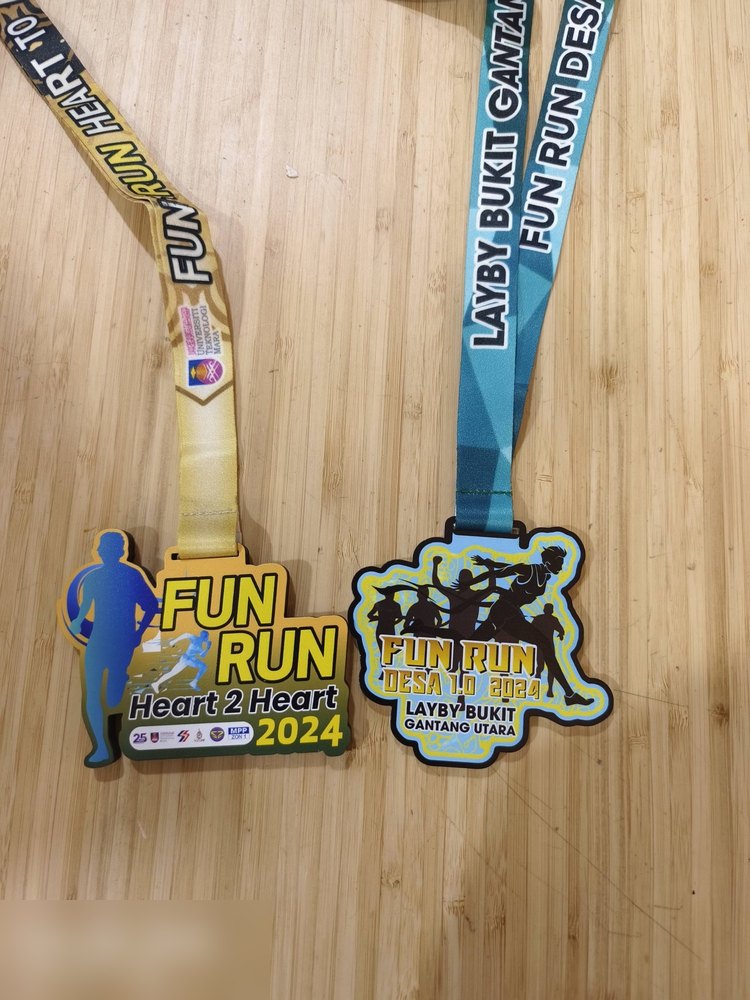 Dua medal fun run bersebelahan: 'FUN RUN Heart 2 Heart 2024' (kuning, silhouette pelari biru) dan 'FUN RUN DESA 1.0 2024 Layby Bukit Gantang Utara' (biru-hijau neon dengan kumpulan pelari). Bagus untu