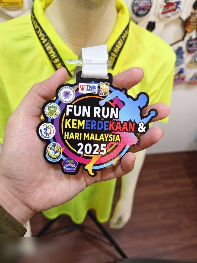 Medal Fun Run Kemerdekaan & Hari Malaysia 2025 anjuran TNB Genco. Bentuk custom, latar hitam dengan silhouette pelari dan pancaran warna biru, merah jambu, kuning. Ada beberapa logo penganjur di sebel