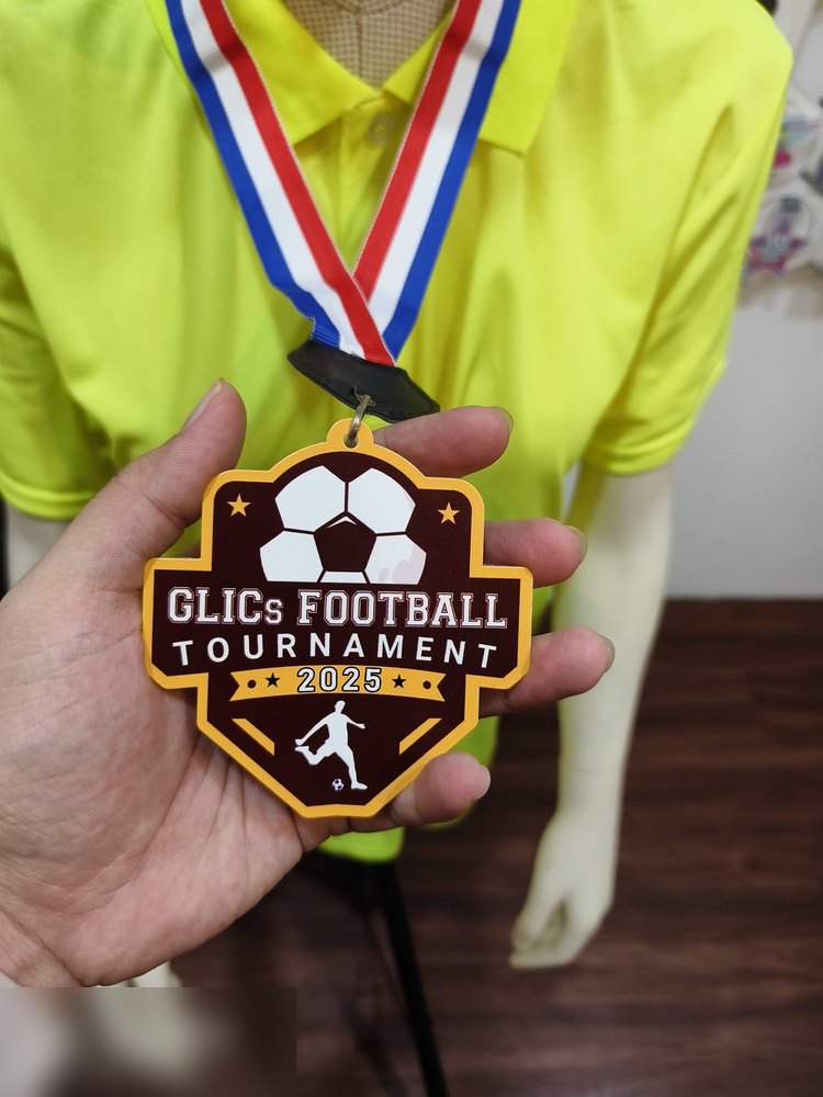Medal GLICs Football Tournament 2025. Bentuk custom perisai dengan bola sepak besar di atas, bintang dan silhouette pemain menendang. Warna marun gelap dengan bingkai kuning emas. Reben jalur merah-pu