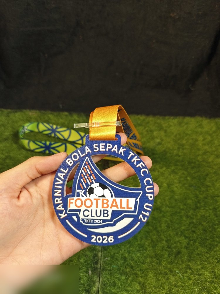 Medal Karnival Bola Sepak TKFC Cup U12 2026. Bentuk bulat biru dengan potongan cut-out segi tiga di tengah, logo TKFC Football Club dan bola sepak. Teks melengkung 'KARNIVAL BOLA SEPAK TKFC CUP U12' d