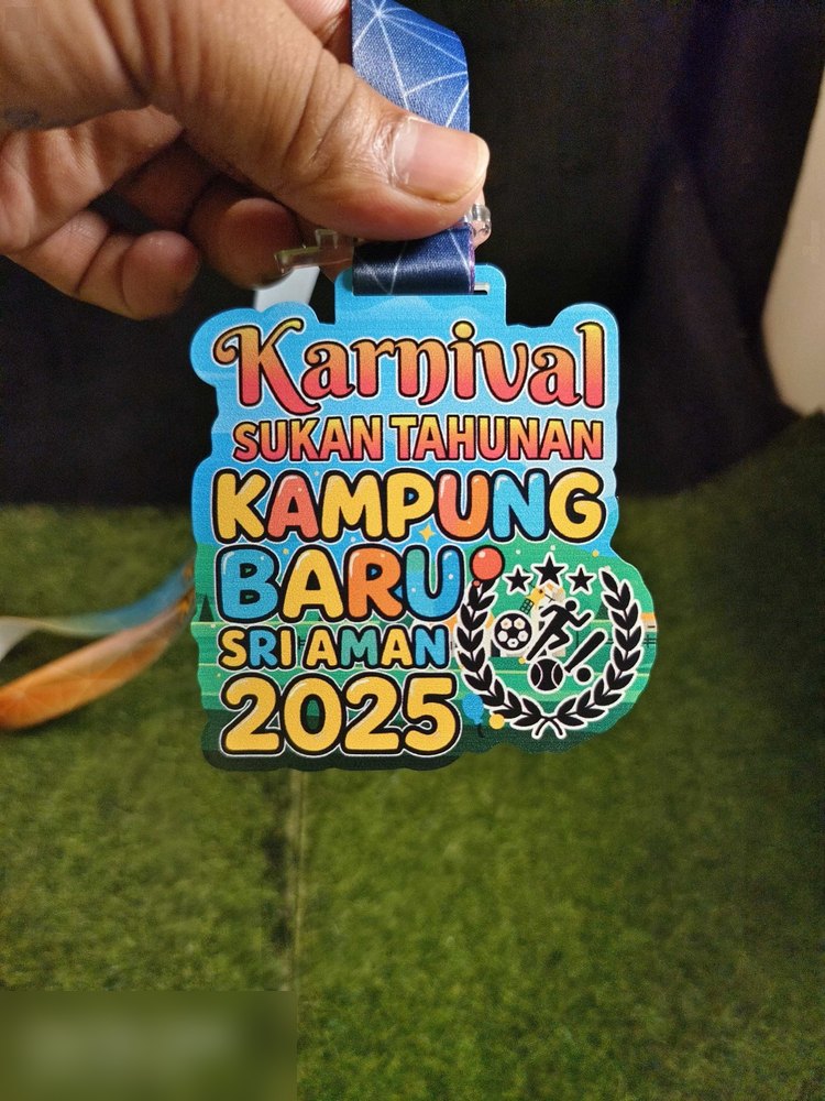 Medal Karnival Sukan Tahunan Kampung Baru Sri Aman 2025 dipegang di tangan — jelas tunjuk teks berwarna-warni bertindan, kalungan daun dan ikon sukan. Sudut berbeza daripada gambar berbaring.