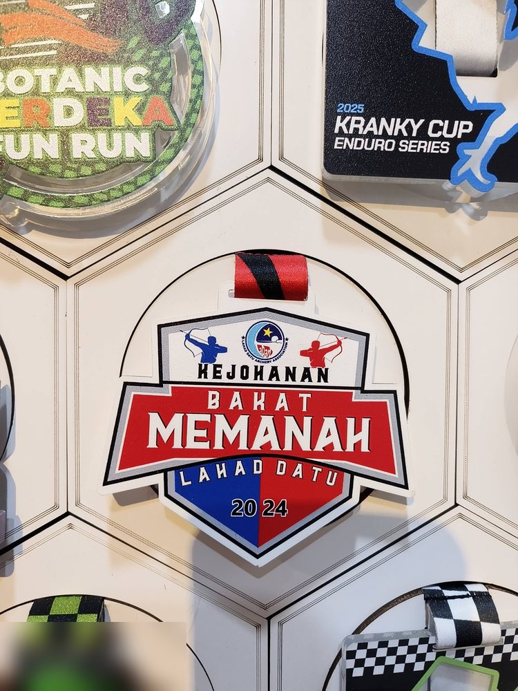 Medal Kejohanan Bakat Memanah Lahad Datu 2024 (Sabah). Bentuk perisai dgn bingkai putih, latar MERAH dan BIRU, dua pemanah biru dan merah di atas. Reben merah-hitam. Dipamerkan di dinding galeri.