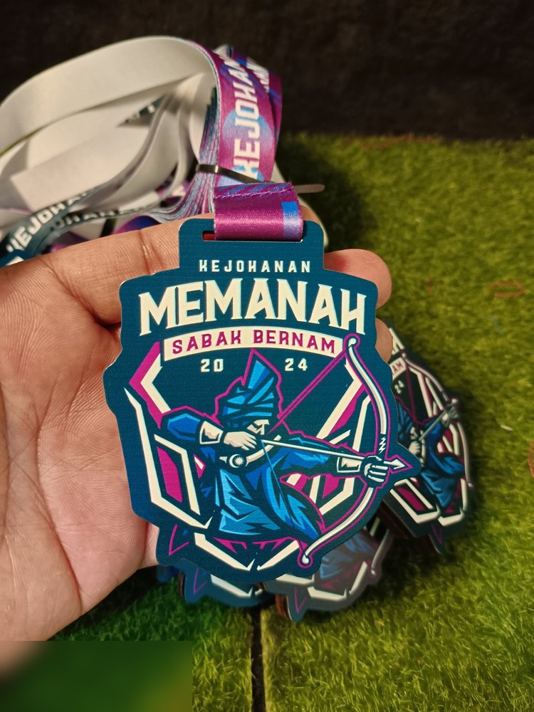 Medal Kejohanan Memanah Sabak Bernam 2024. Bentuk custom, warna teal/biru petrol dgn pemanah dan corak merah jambu terang. Lanyard ungu-biru bercetak. Versi 2024 (bandingkan dgn versi 2025 yang ungu).