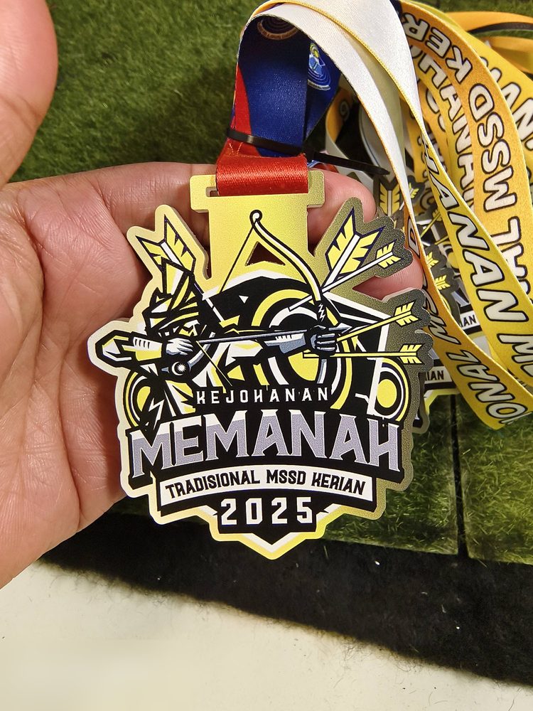 Medal memanah MSSD Kerian 2025 — sama design dengan Image 1, tetapi gambar ini tunjuk lanyard kuning bercetak penuh 'KEJOHANAN MEMANAH TRADISIONAL MSSD KERIAN' dan reben merah di clip. Bagus untuk tun