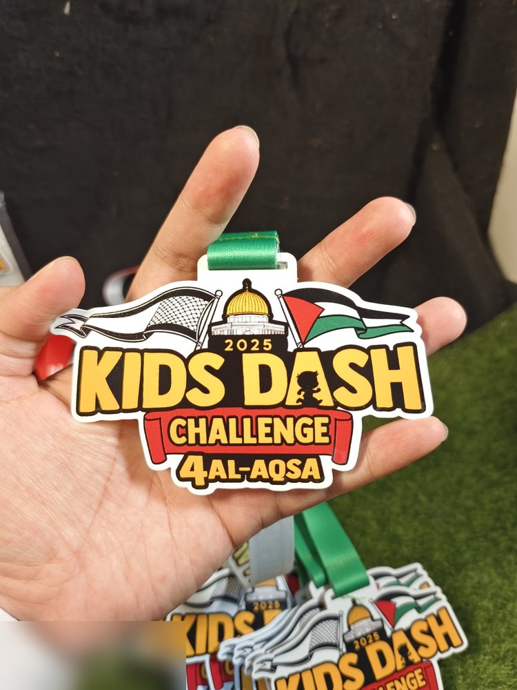 Medal Kids Dash Challenge 4 Al-Aqsa 2025 dipegang di tangan — jelas tunjuk kubah emas Masjid Al-Aqsa, bendera Palestin, kaffiyeh dan teks kuning. Reben hijau. Sudut berbeza daripada gambar pukal.