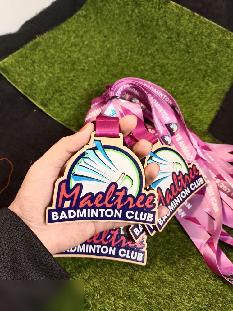Medal Maeltree Badminton Club. Bentuk custom melengkung seperti kubah, latar biru navy dgn bulu tangkis putih-biru memancar dan hijau. Nama 'Maeltree' dlm tulisan sambung merah. Lanyard merah jambu be