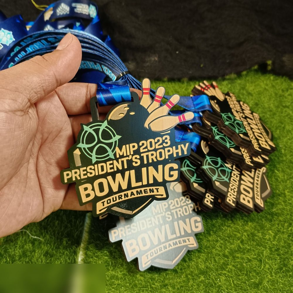 Medal MIP 2023 President's Trophy Bowling Tournament. Bentuk custom dengan pin bowling dan bola timbul di bahagian atas. Warna hijau gelap/hitam dgn teks emas dan logo hijau mint. Gambar tunjuk DUA ve