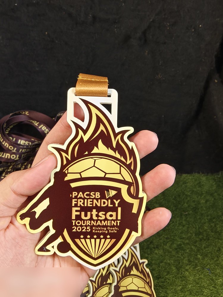 Medal PACSB Friendly Futsal Tournament 2025 diambil dekat — jelas tunjuk kualiti cetakan warna marun dan emas serta bentuk perisai berapi. Ada beberapa keping di bawah.