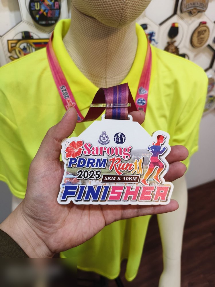 Medal Sarong PDRM Run 2025 Finisher, kategori 5KM & 10KM. Bentuk custom, latar putih dgn BUNGA RAYA merah, pelari merah jambu, logo PDRM dan gambar latar bandar. Lanyard merah jambu bercetak. Contoh m