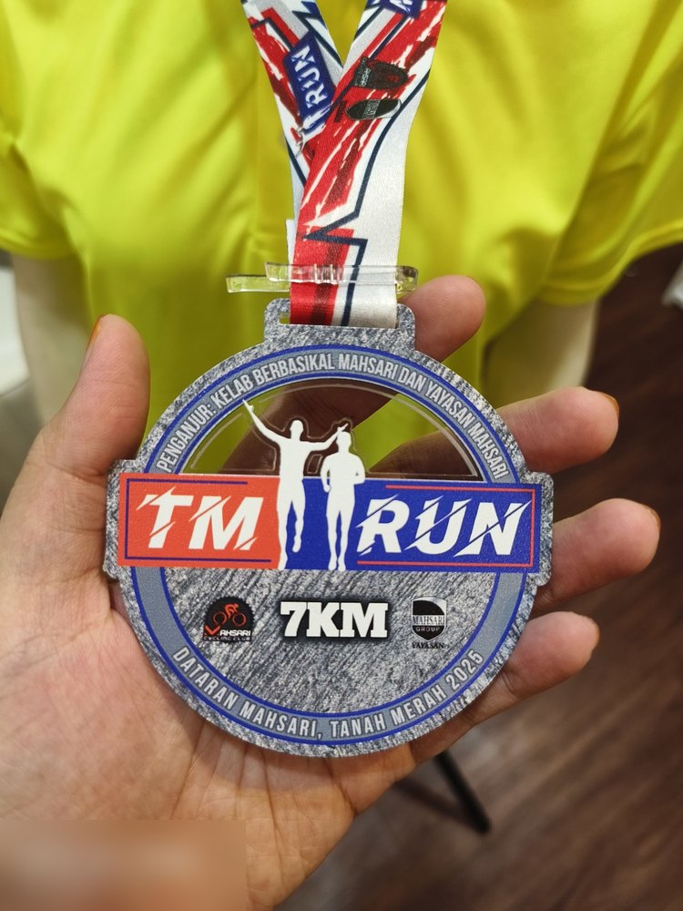Medal larian TM RUN 7KM 2025, anjuran Kelab Berbasikal Mahsari dan Yayasan Mahsari di Dataran Mahsari, Tanah Merah. Bentuk bulat dengan tekstur kelabu marble, teks 'TM RUN' merah-biru dan dua silhouet